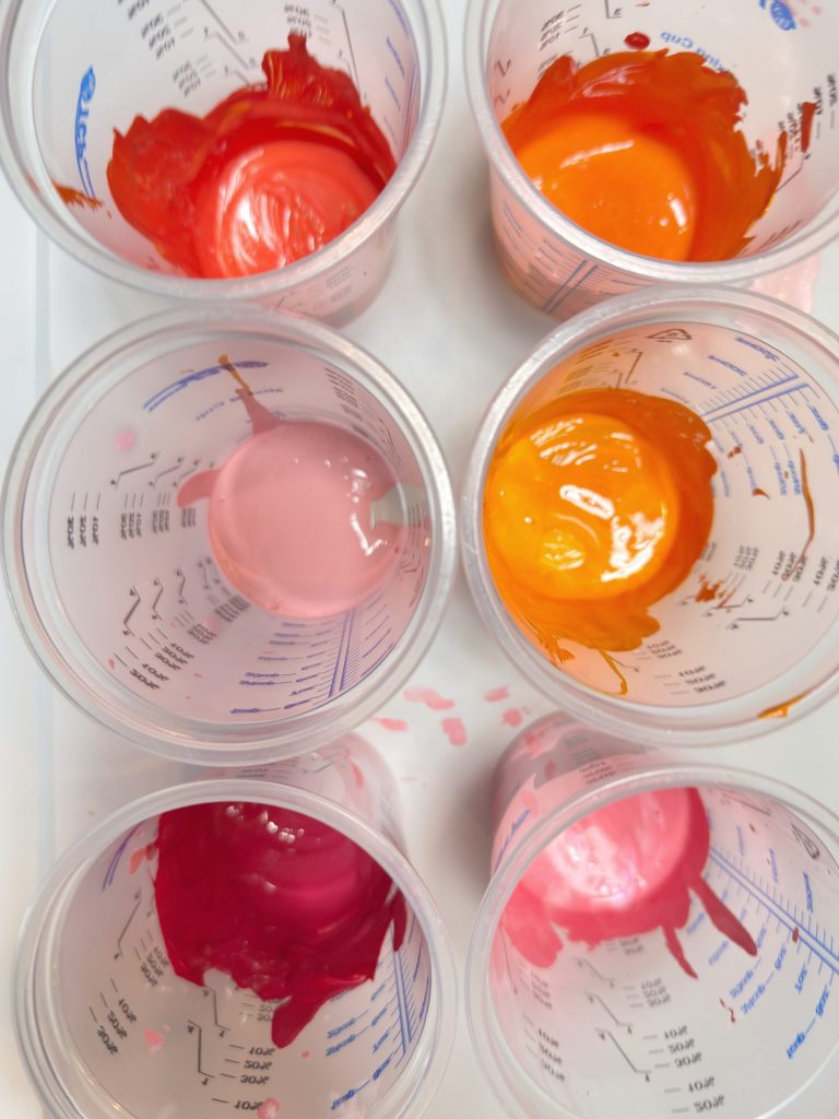 Six transparent plastic cups containing various shades of red, orange, and pink paint are arranged in two rows on a white surface.