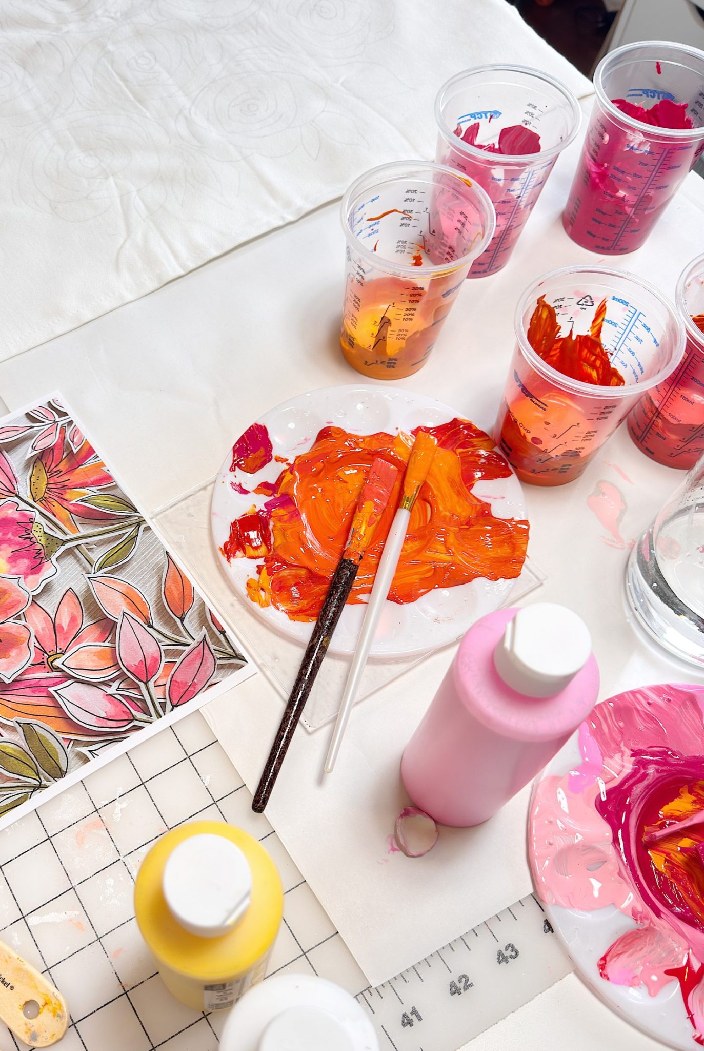 Paintbrushes and a plate with orange and red paint, paint bottles, cups with mixed colors, and a floral artwork sit on a white workspace.