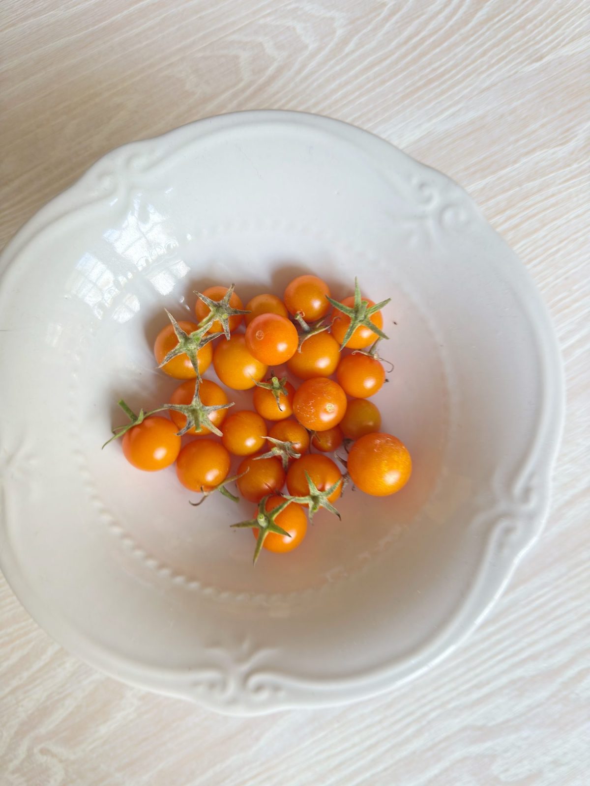 A white ceramic bowl contains several small orange cherry tomatoes with green stems, placed on a light wood surface.