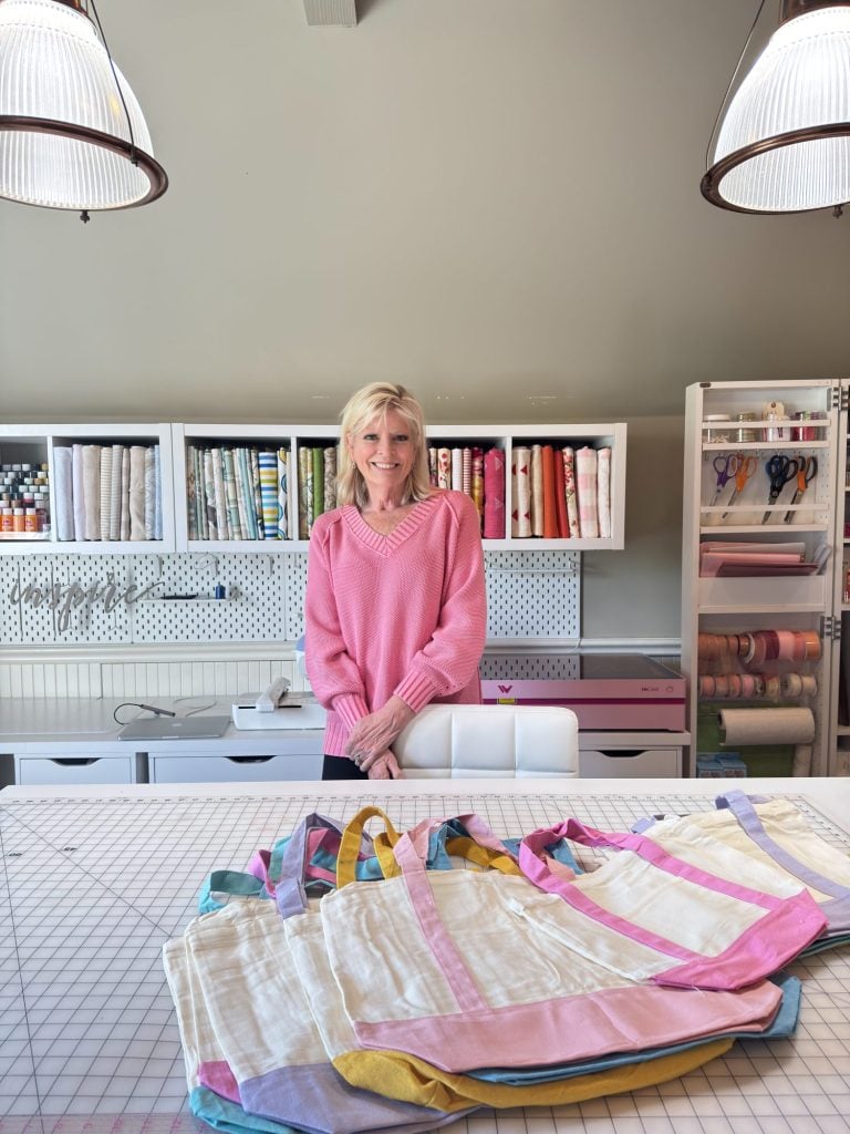 A woman in a pink sweater stands behind a table with several colorful tote bags in a well-organized fabric and craft room.