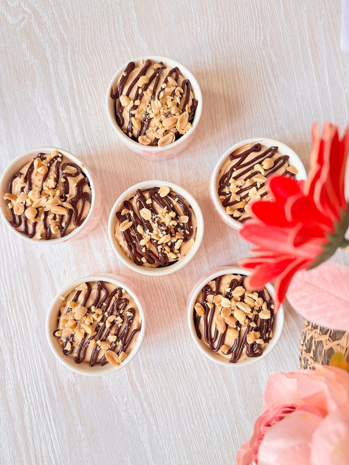 Six small bowls filled with a dessert topped with chocolate drizzle and chopped nuts sit on a light wooden surface next to red and pink flowers.