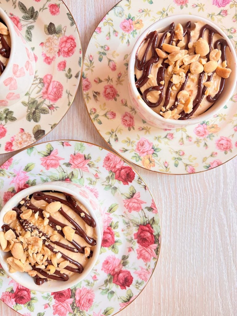 Three floral-patterned saucers with matching cups filled with dessert topped with chocolate drizzle and chopped nuts on a light wooden surface.