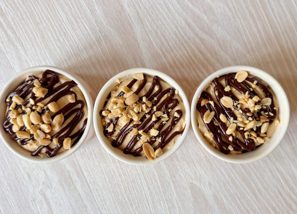 Three small bowls filled with a creamy dessert, topped with chocolate drizzle and chopped peanuts, arranged in a row on a light wooden surface.