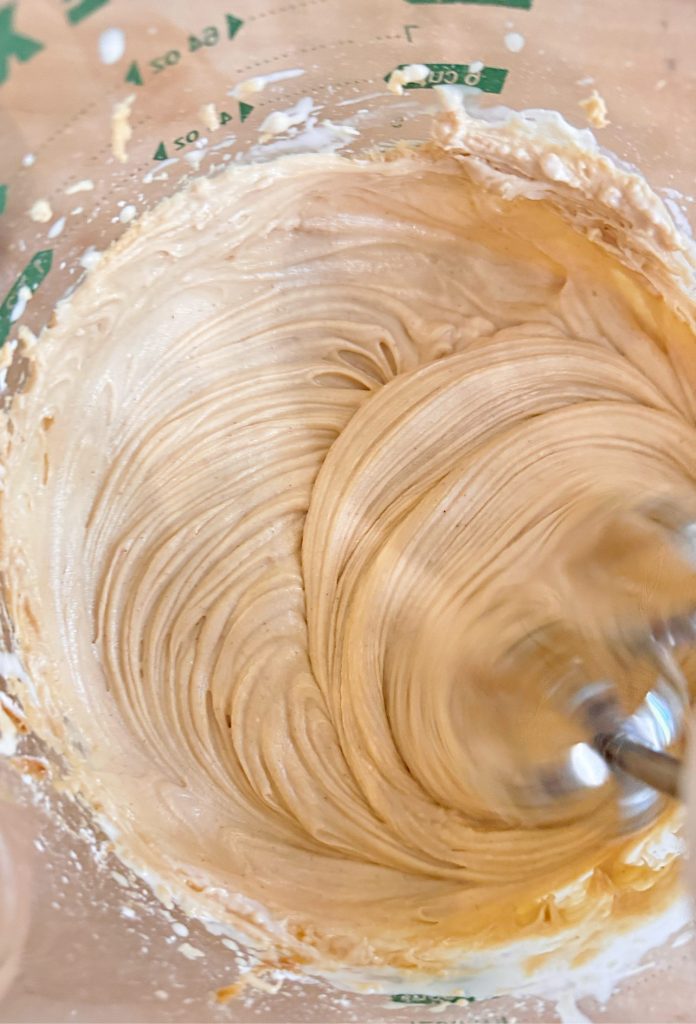 Close-up of a mixing bowl with creamy, light brown batter being blended by an electric mixer. Measurement markings are visible on the side of the bowl.