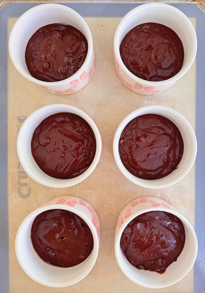 Six white ramekins filled with chocolate cake batter are arranged on a silicone baking mat, ready to be baked.