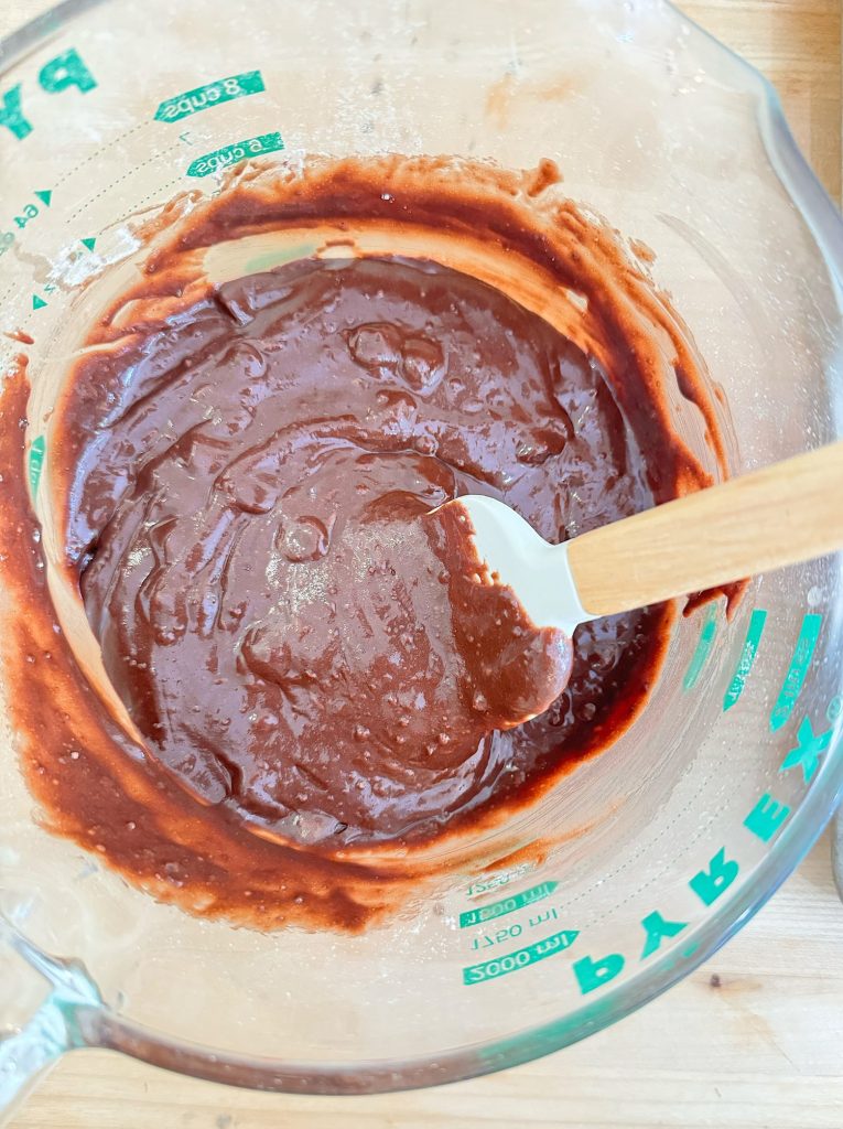 A glass mixing bowl with chocolate batter and a white silicone spatula on a wooden surface.