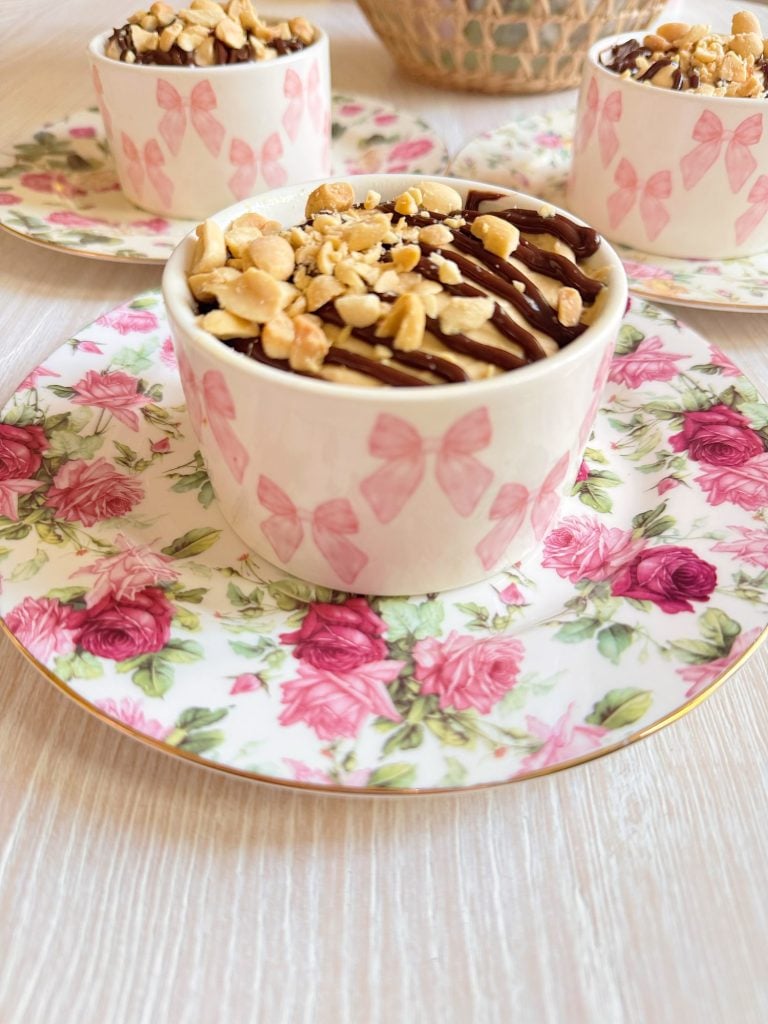 A ceramic cup with pink butterfly designs, filled with dessert topped with chopped nuts and chocolate drizzle, sits on a floral-patterned saucer.