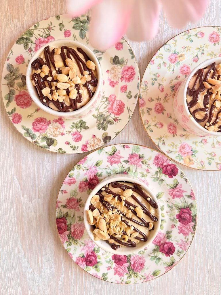 Three small bowls of dessert topped with chocolate sauce and peanuts, placed on floral-patterned plates on a light wooden surface.
