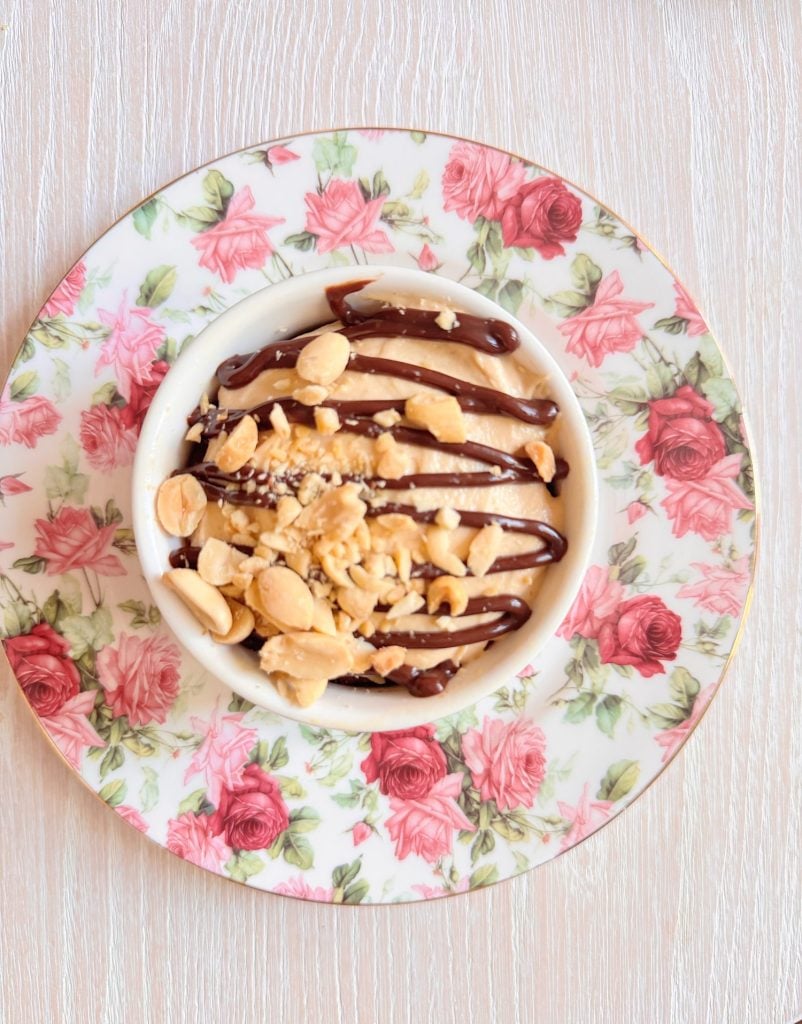 A bowl of dessert topped with chocolate sauce and crushed peanuts, served on a floral-patterned plate on a light wooden surface.