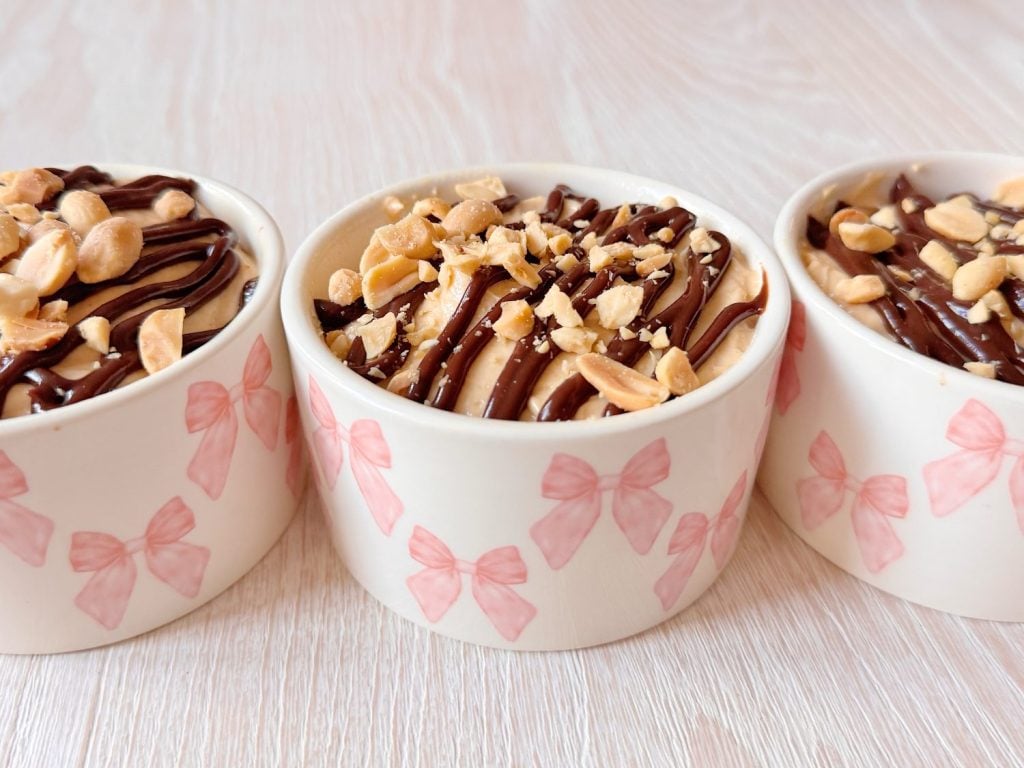 Three small cups with pink bow designs filled with dessert, topped with chocolate drizzle and chopped peanuts, on a light wooden surface.