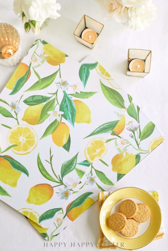 A tabletop with a lemon-patterned placemat, a yellow plate with cookies, two lit candles in gold holders, and white flowers in the background.
