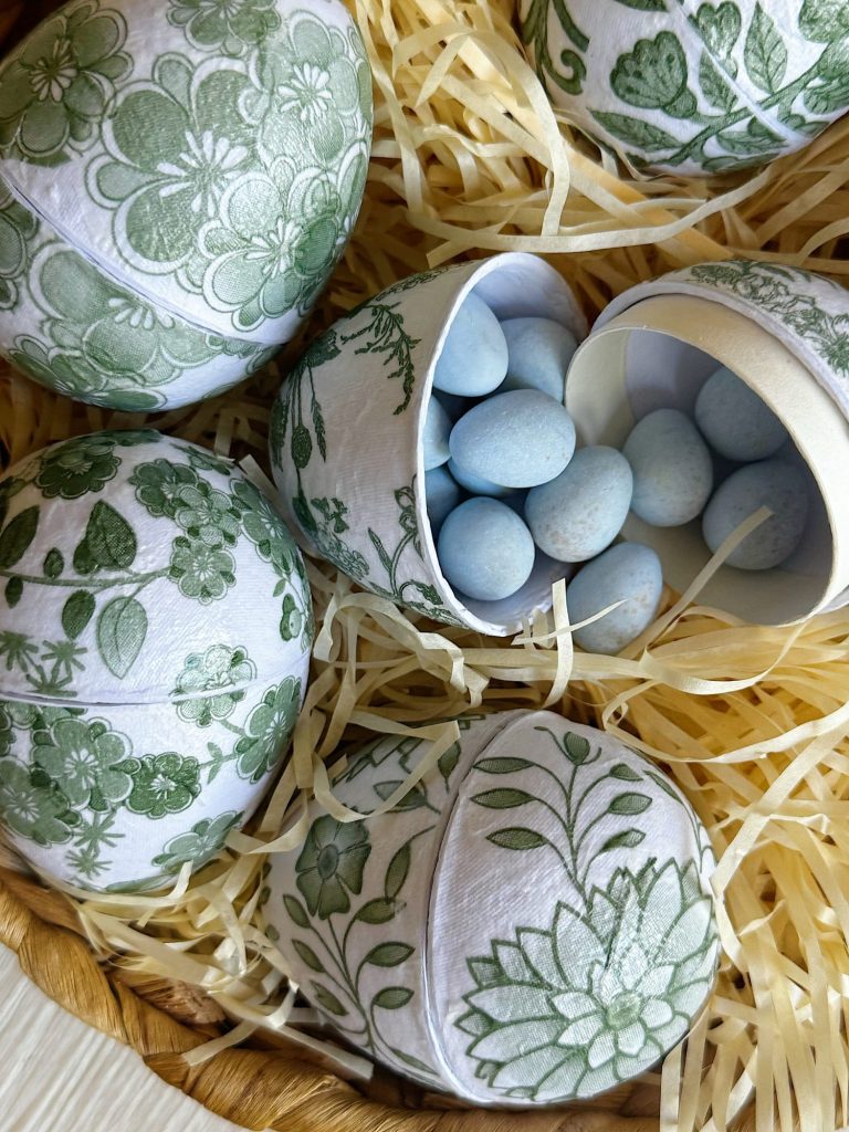 Decorative eggs with green floral patterns sit in straw, one egg is opened to reveal blue candy eggs inside.