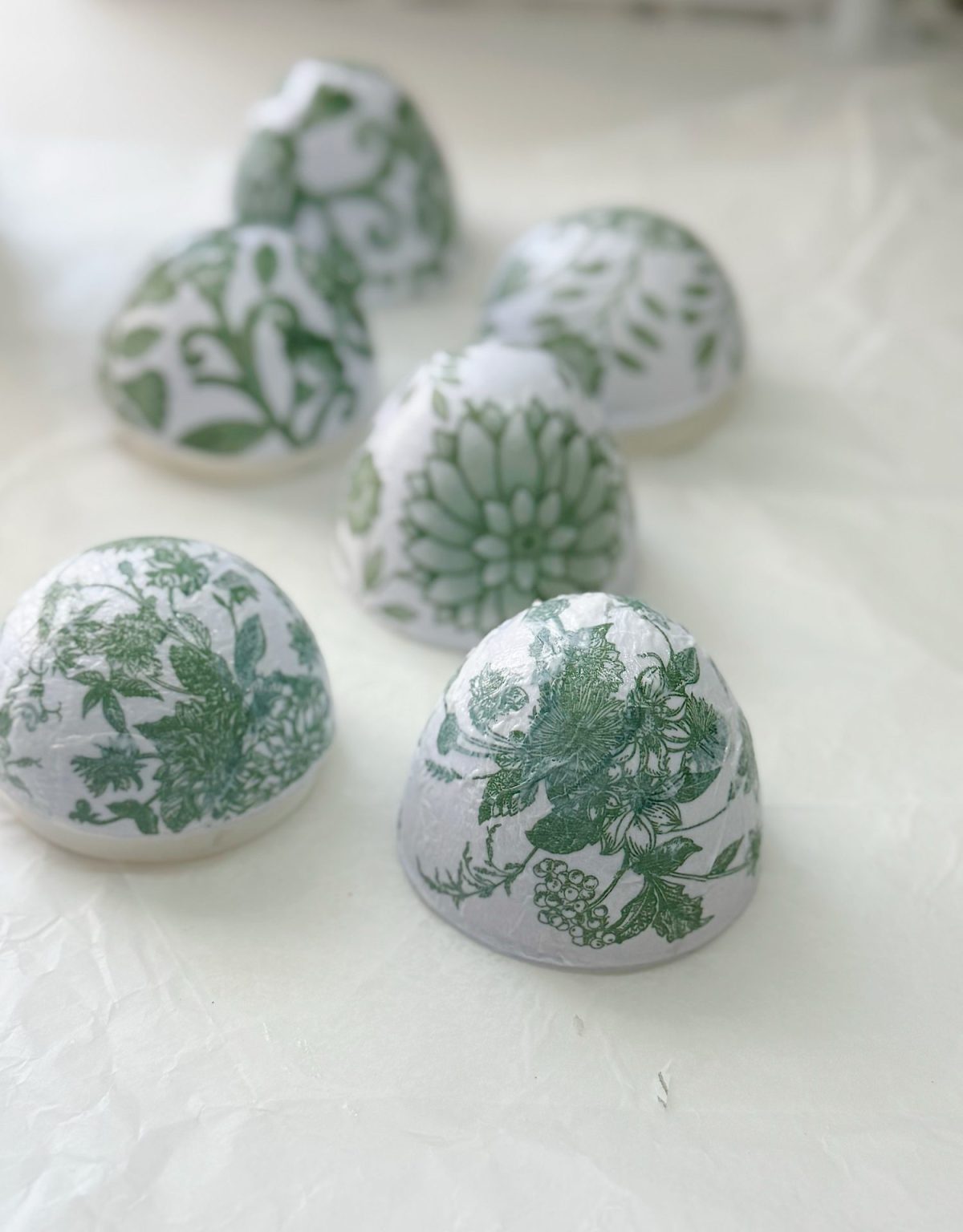 Six dome-shaped objects with green floral patterns on a white background are arranged on a crinkled white surface.