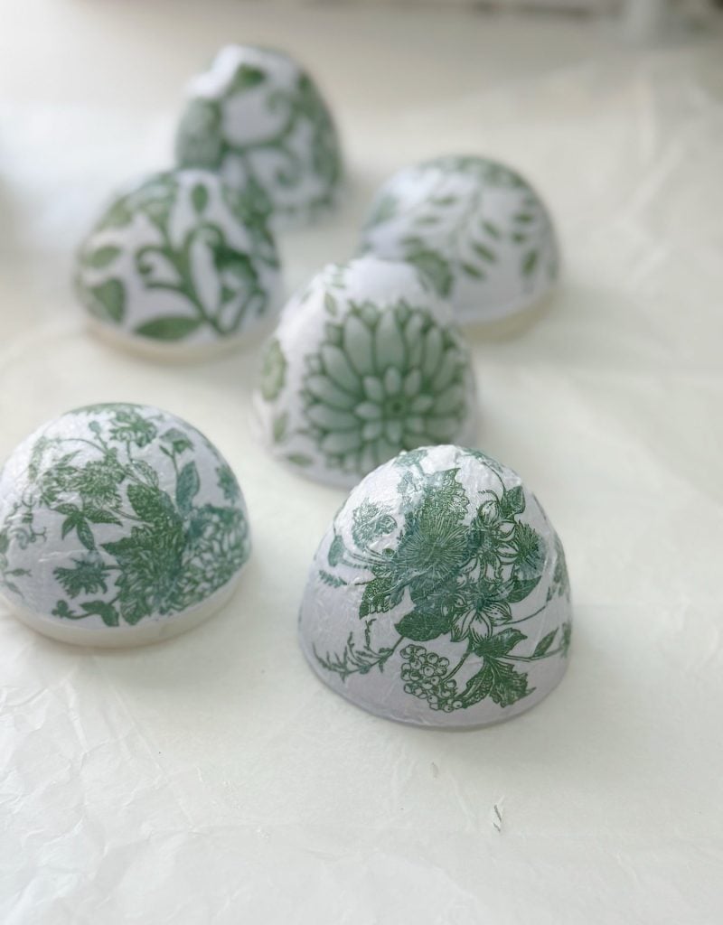 Six dome-shaped objects with green floral patterns on a white background are arranged on a crinkled white surface.