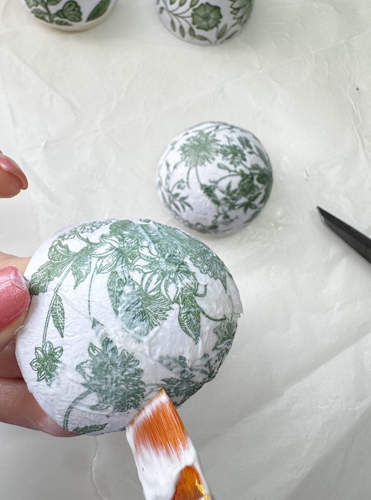 A hand applies glue to a round object covered with green and white floral-patterned paper, with scissors and another decorated object nearby on a white surface.