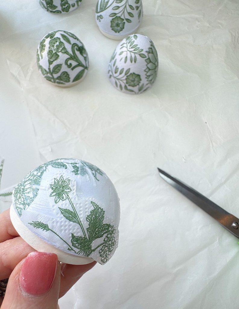 Hand holding a white egg decorated with green floral patterns, with more decorated eggs, a pair of scissors, and paper visible in the background.