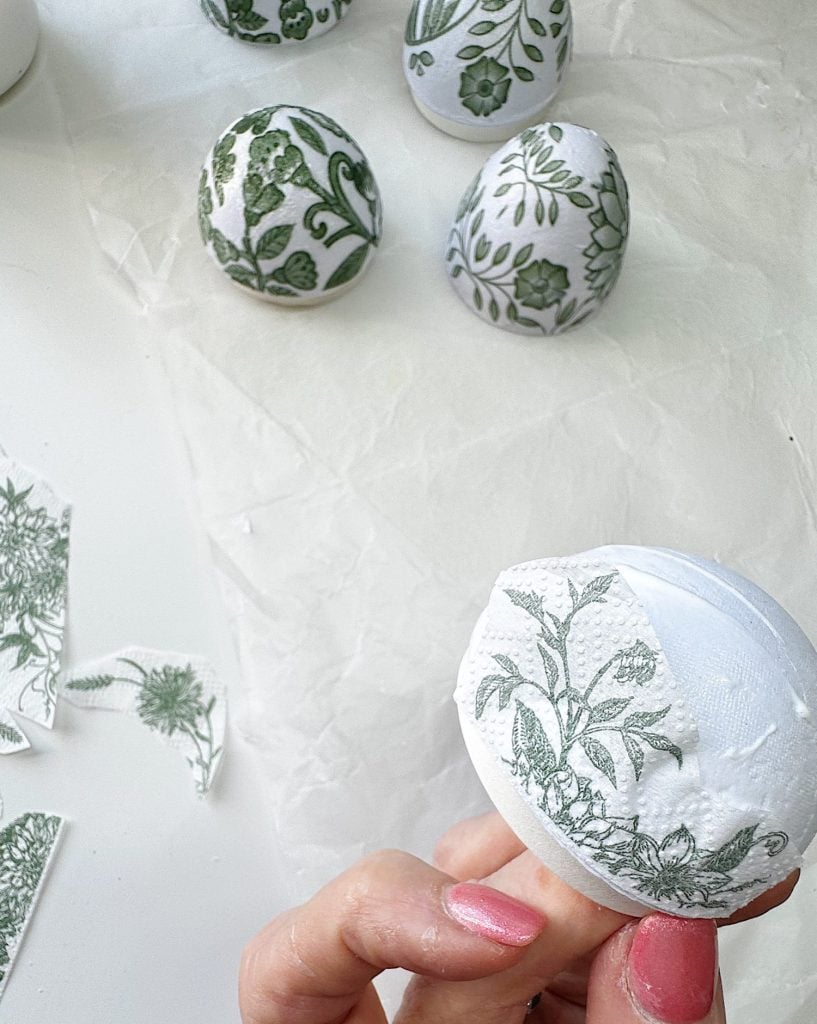 A hand decorates an egg with a green floral-patterned paper; three finished eggs with similar designs are on white paper in the background.