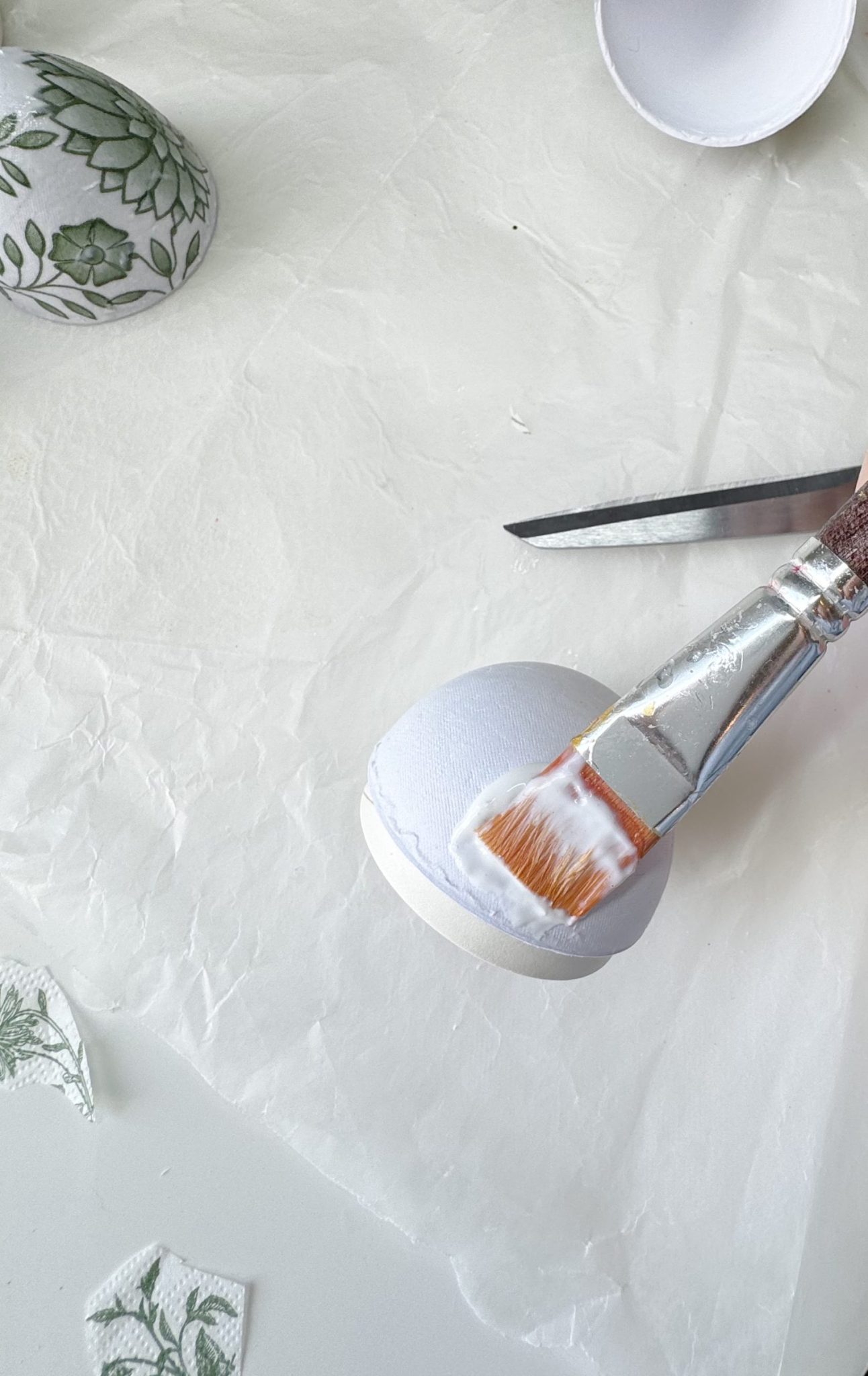 A paintbrush applies glue to a white eggshell half on parchment paper; scissors and decoupage paper with green patterns are nearby.