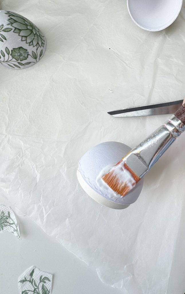 A paintbrush applies glue to a white eggshell half on parchment paper; scissors and decoupage paper with green patterns are nearby.
