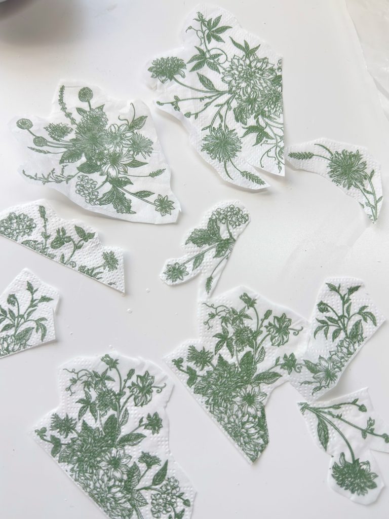 Several pieces of white paper napkin with green floral patterns are cut into irregular shapes and spread out on a white surface.