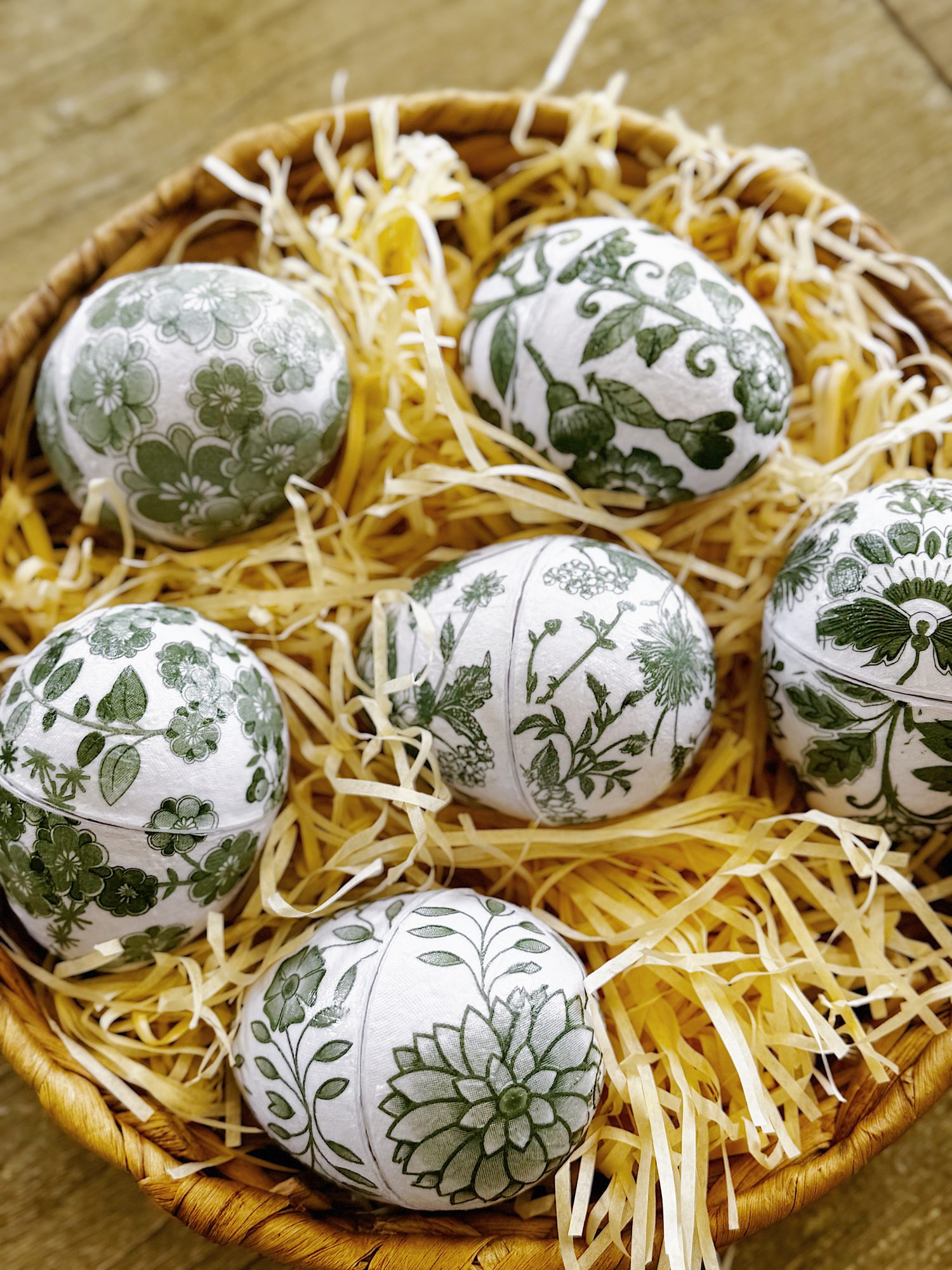 A basket filled with six white eggs decorated with green floral and botanical patterns, resting on yellow straw.