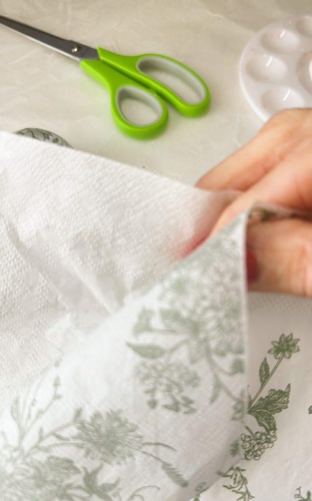 A hand separates two layers of a floral-printed paper napkin. Green scissors and a white palette are visible in the background.