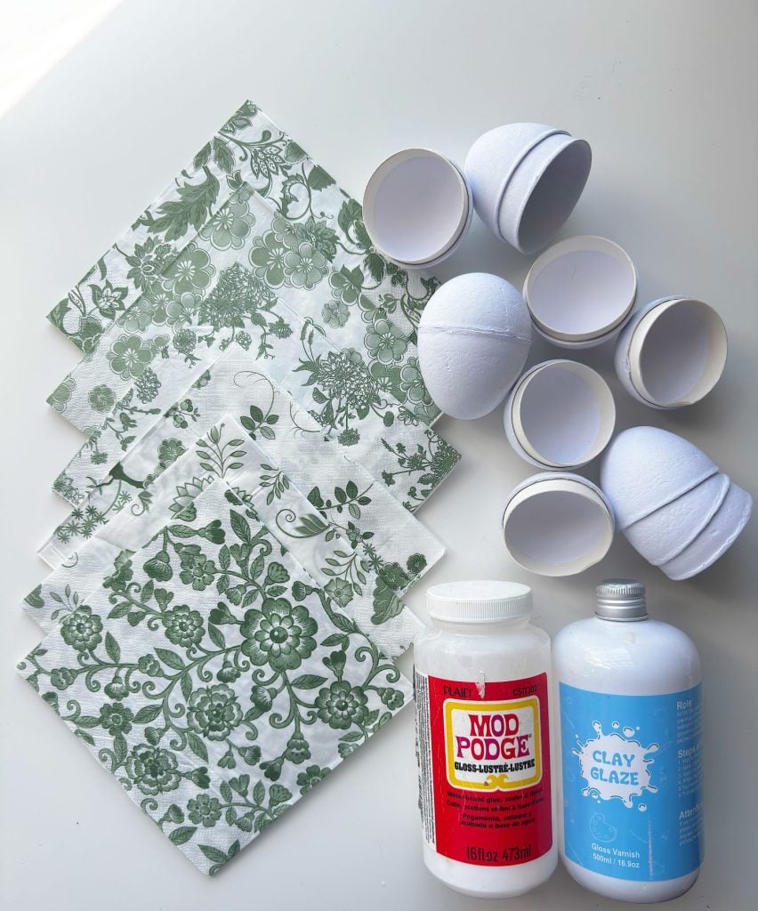 Green floral paper napkins, white split plastic eggs, a bottle of Mod Podge, and a bottle of clay glaze are arranged on a white surface for a craft project.
