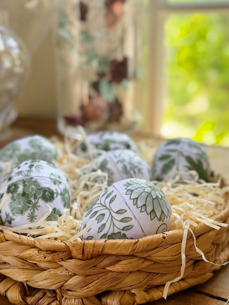 A woven basket filled with decorative eggs featuring green floral patterns rests on straw, placed near a window with sunlight coming through.