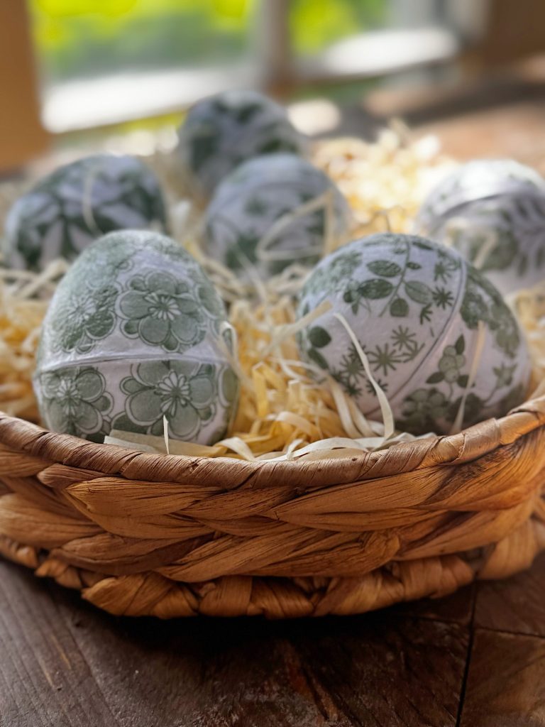 A woven basket filled with decorative eggs featuring green floral patterns, resting on a bed of straw on a wooden surface near a window.