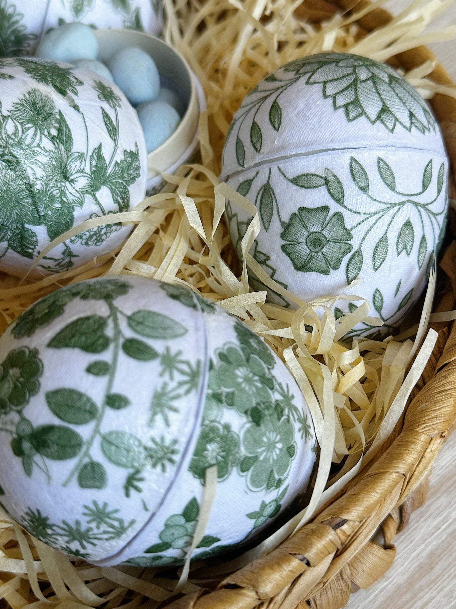 Close-up of large eggs decorated with green floral patterns, placed in a basket filled with straw, with small blue eggs in the background.