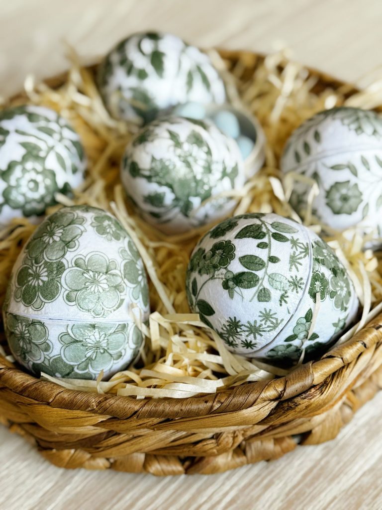 A woven basket filled with six eggs decorated with green floral patterns, resting on a bed of straw.