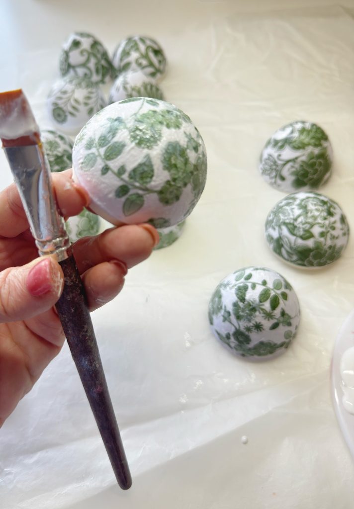 A hand holds a paintbrush and a round object decorated with green leaf patterns; several similar decorated objects are on a white surface in the background.