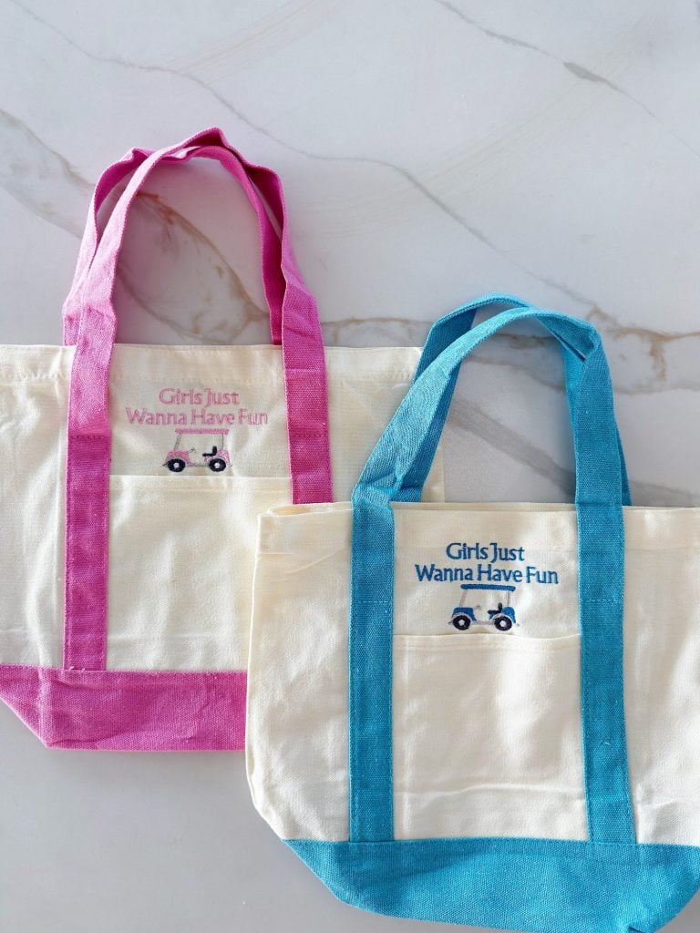 Two canvas tote bags with colored handles, one pink and one blue, feature a small car graphic and the phrase "Girls Just Wanna Have Fun" on a white marble surface.