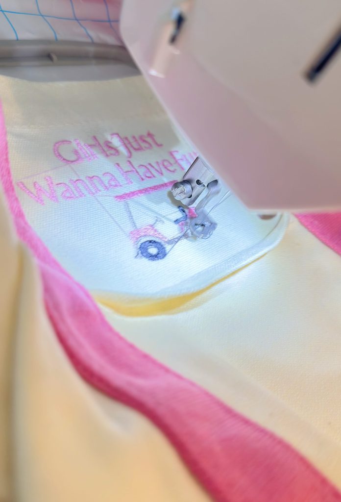 A sewing machine stitching the phrase "Girls Just Wanna Have Fun" in pink thread onto white fabric with pink trim.