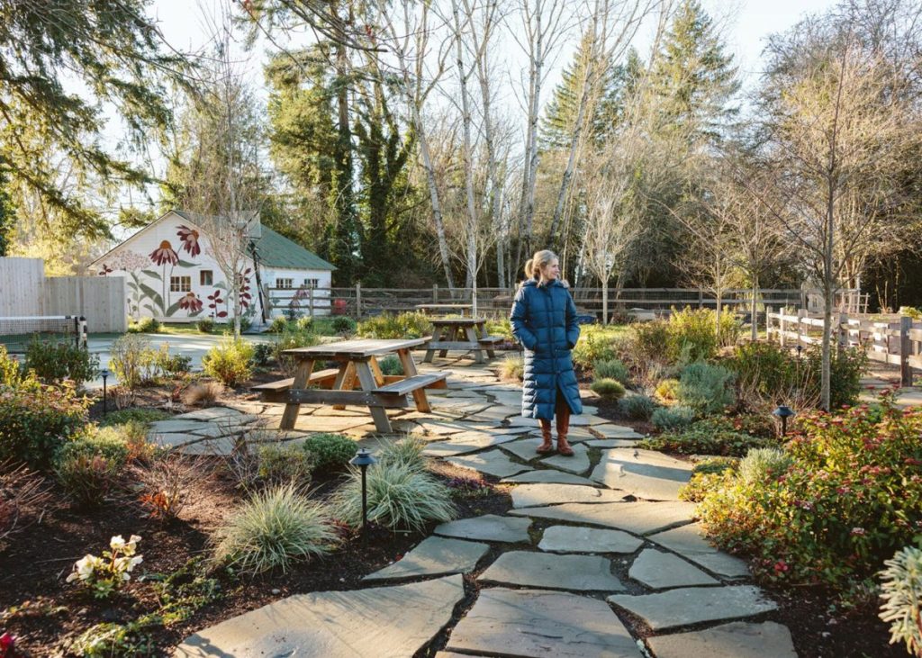 A person in a blue coat stands on a stone path in a landscaped garden with picnic tables, surrounded by plants and trees, near a white building.