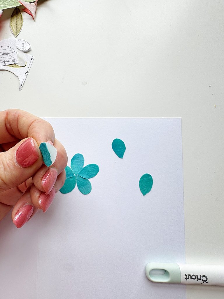 A hand with pink nails holds a blue paper petal over a white sheet with more paper petals and a Cricut tool nearby.