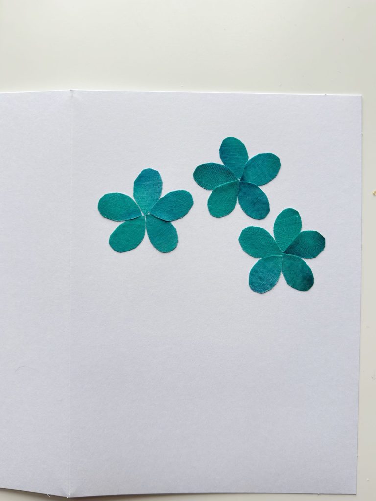Three teal paper flower shapes are arranged on the right side of an open white card placed on a white surface.