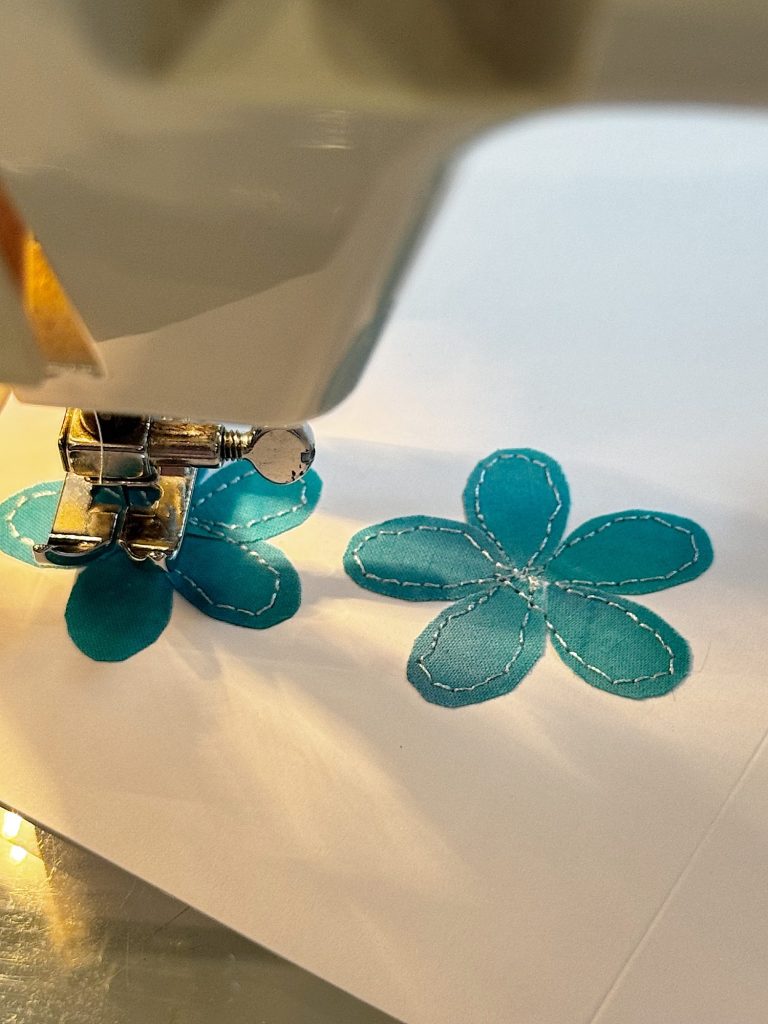 Close-up of a sewing machine stitching blue fabric flower shapes onto a white surface.