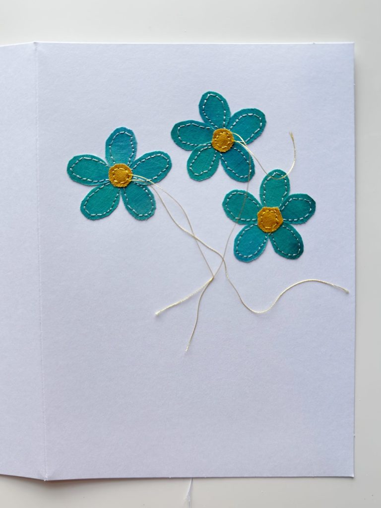 A white card with three blue fabric flowers, each with a yellow center and white thread stems, attached to the right side of the card.