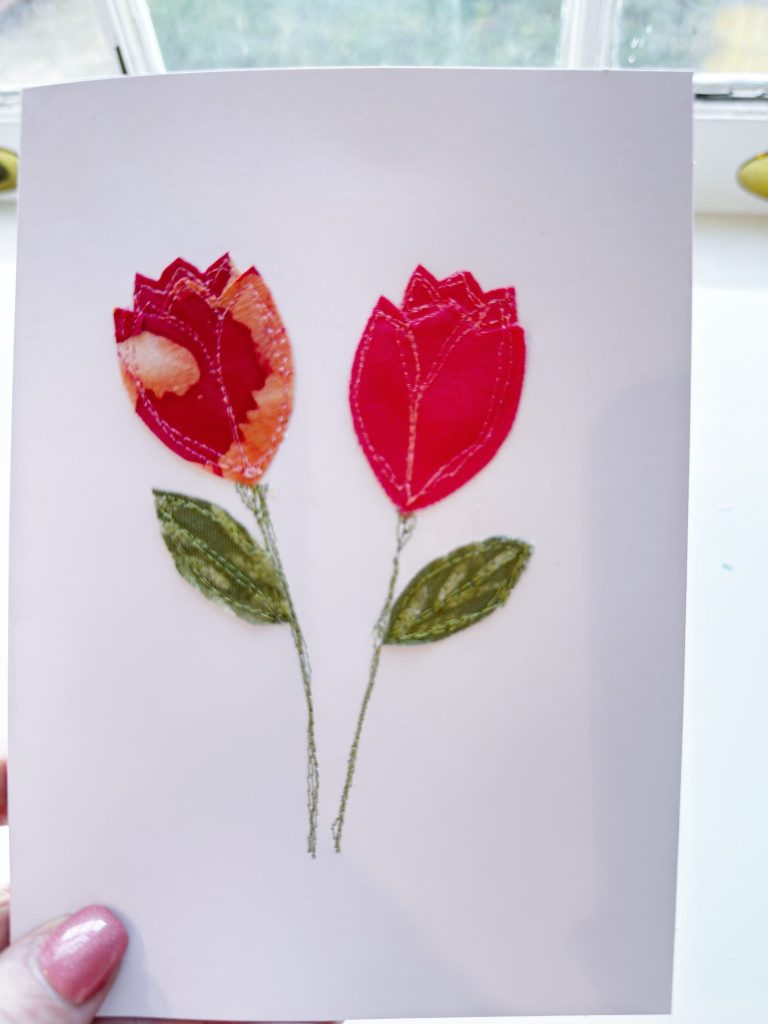 A white card featuring two red fabric tulips with green stitched stems and leaves, held up in front of a window.