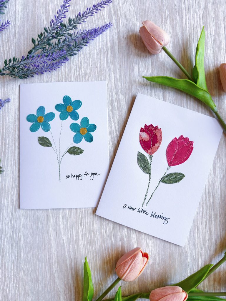 Two greeting cards with hand-drawn flowers, one with blue flowers reads "so happy for you" and one with pink flowers reads "a new little blessing," surrounded by real flowers.
