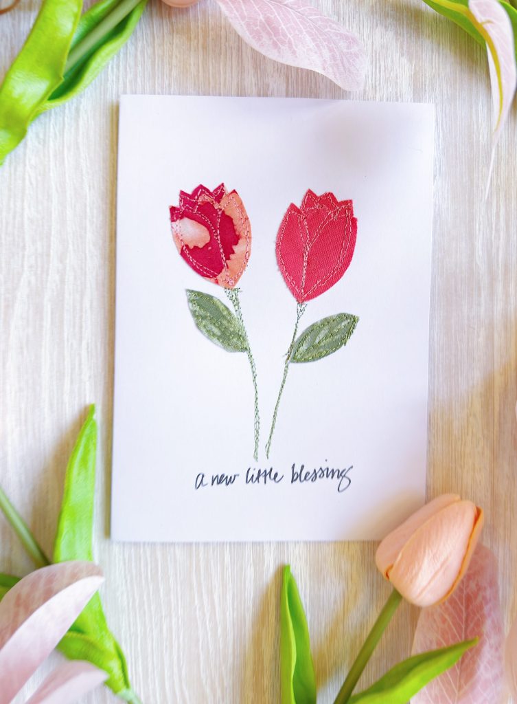 A white card with two red tulip illustrations and the text "a new little blessing," surrounded by assorted artificial flowers on a light wooden surface.