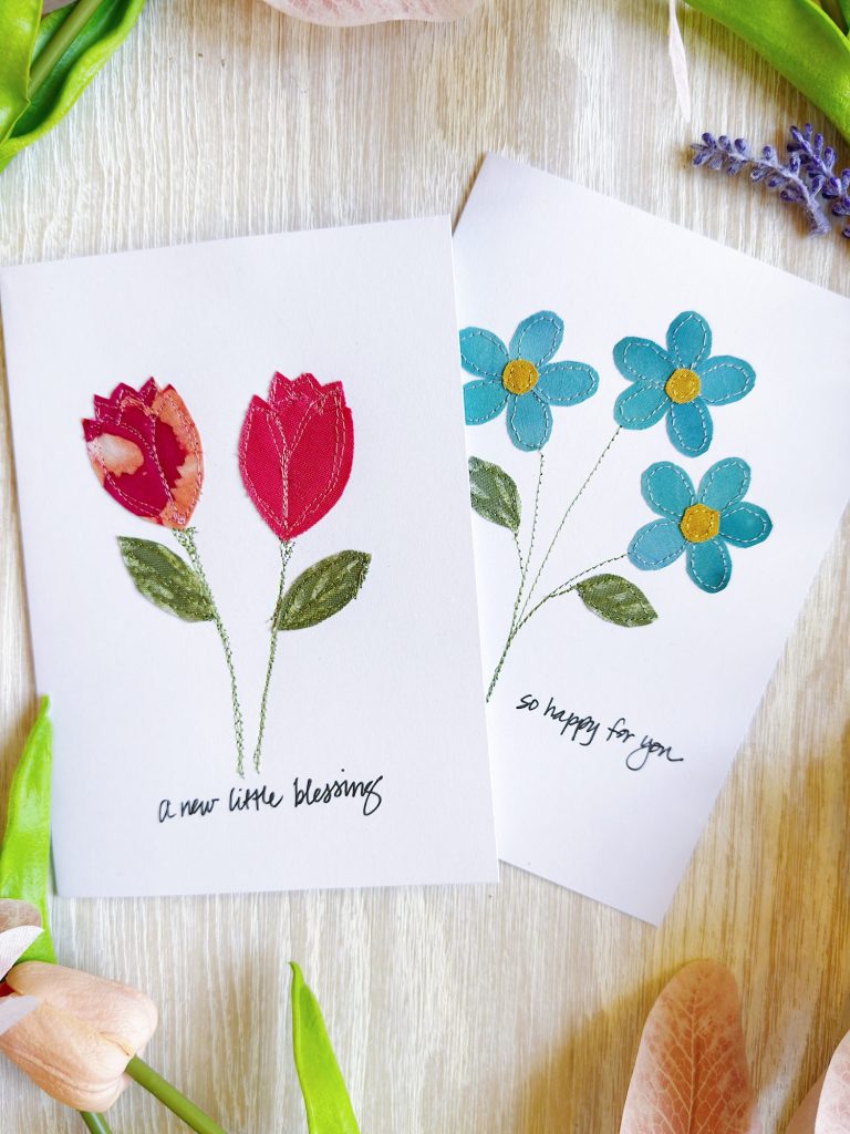 Two handmade greeting cards featuring fabric flower designs and handwritten messages, surrounded by assorted artificial flowers on a light wooden surface.