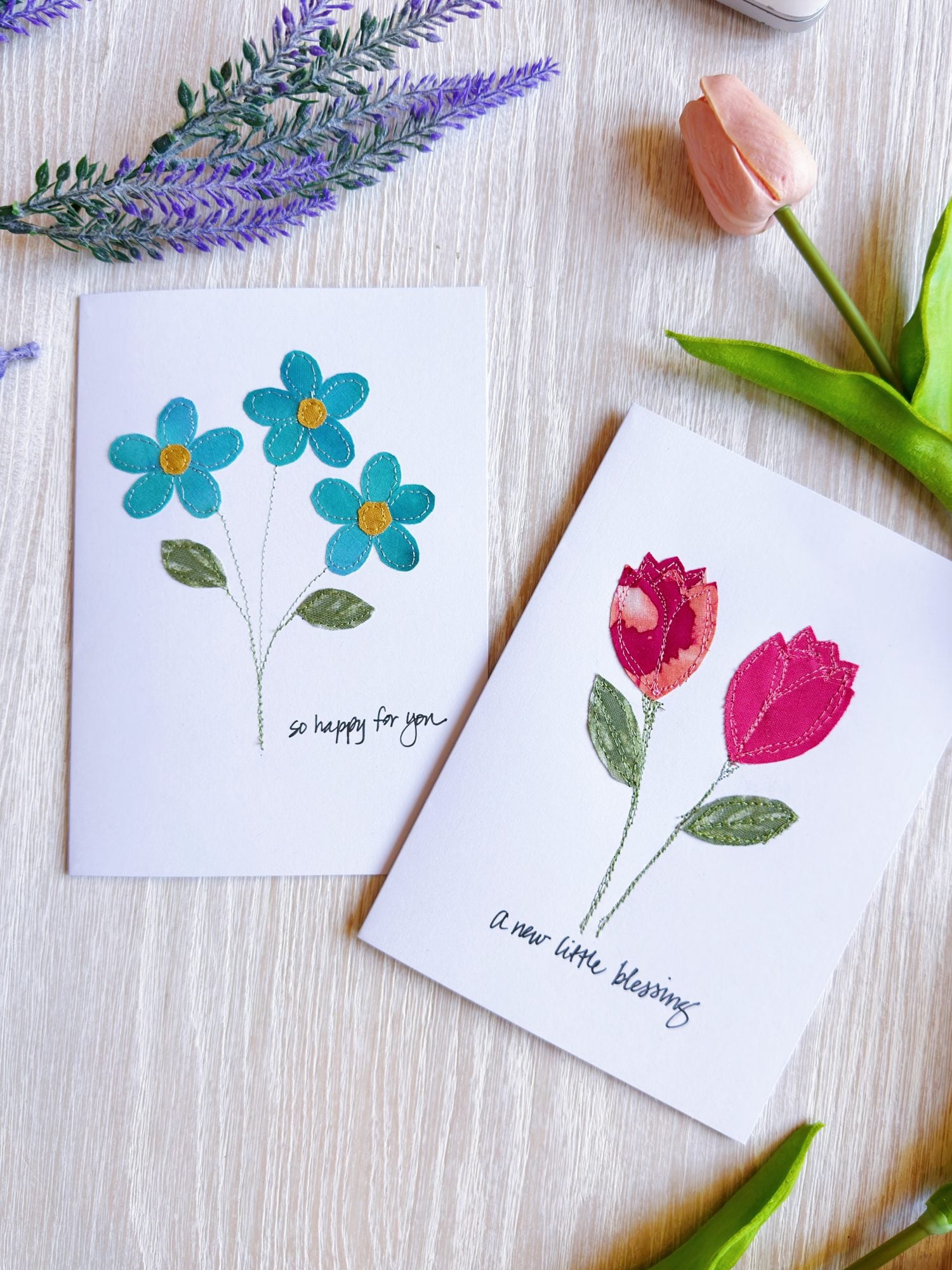 Two greeting cards on a table, each with fabric flower designs and handwritten messages; one says "so happy for you" and the other says "a new little blessing." Flowers and leaves surround them.