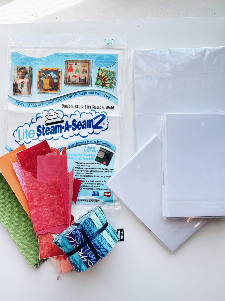 A pack of Lite Steam-A-Seam 2 fusible web, assorted colorful fabric strips, and stacks of white sheets are arranged on a white surface.