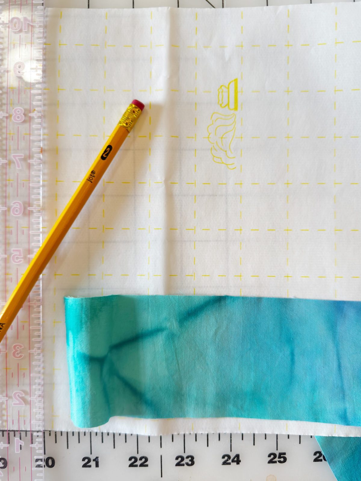 A yellow pencil, a transparent ruler, and a strip of blue-green fabric rest on a gridded cutting mat with markings and a faint embroidery outline on white fabric.
