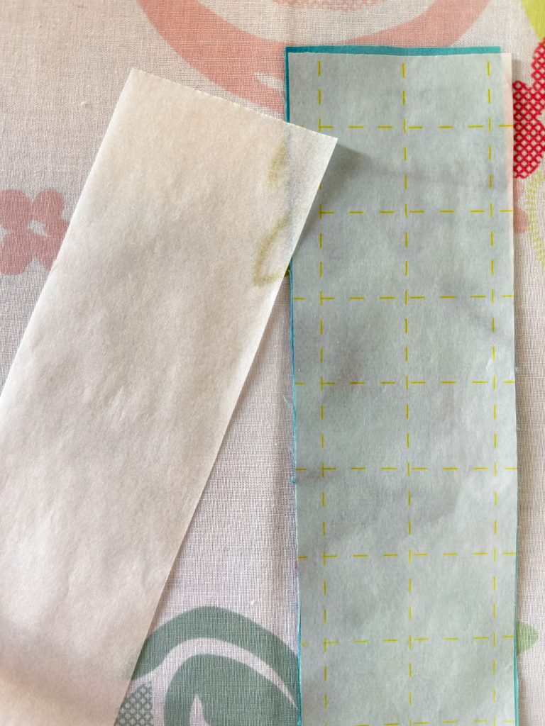 Two strips of paper, one plain and one with a grid printed in yellow, are placed side by side on a patterned fabric surface.