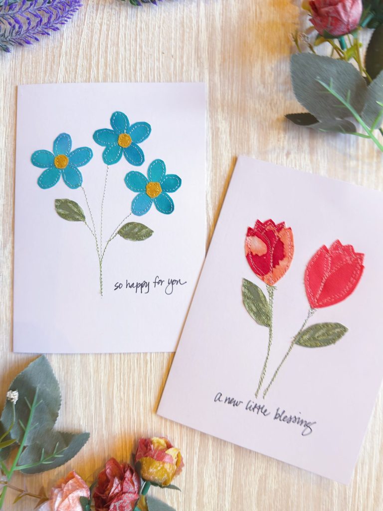 Two greeting cards with fabric flower designs and handwritten text; one says "so happy for you" with blue flowers, the other "a new little blessing" with red flowers.