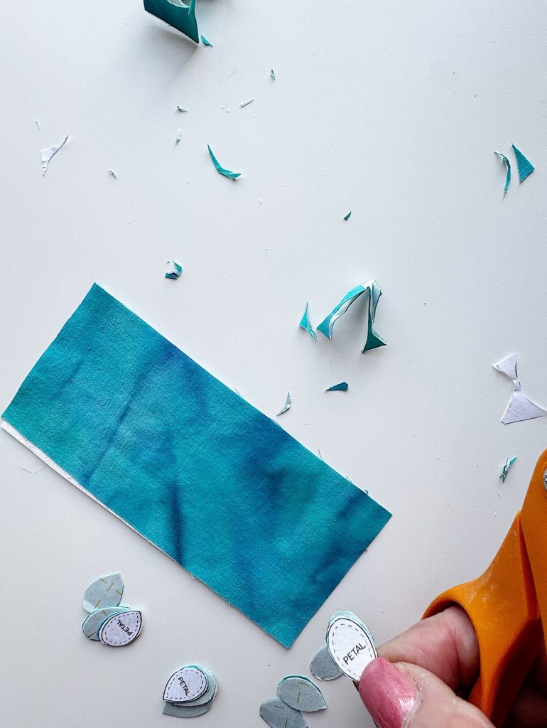 Person cutting out small paper templates labeled "PETAL" with orange scissors beside a rectangular piece of teal fabric and scattered paper scraps on a white surface.