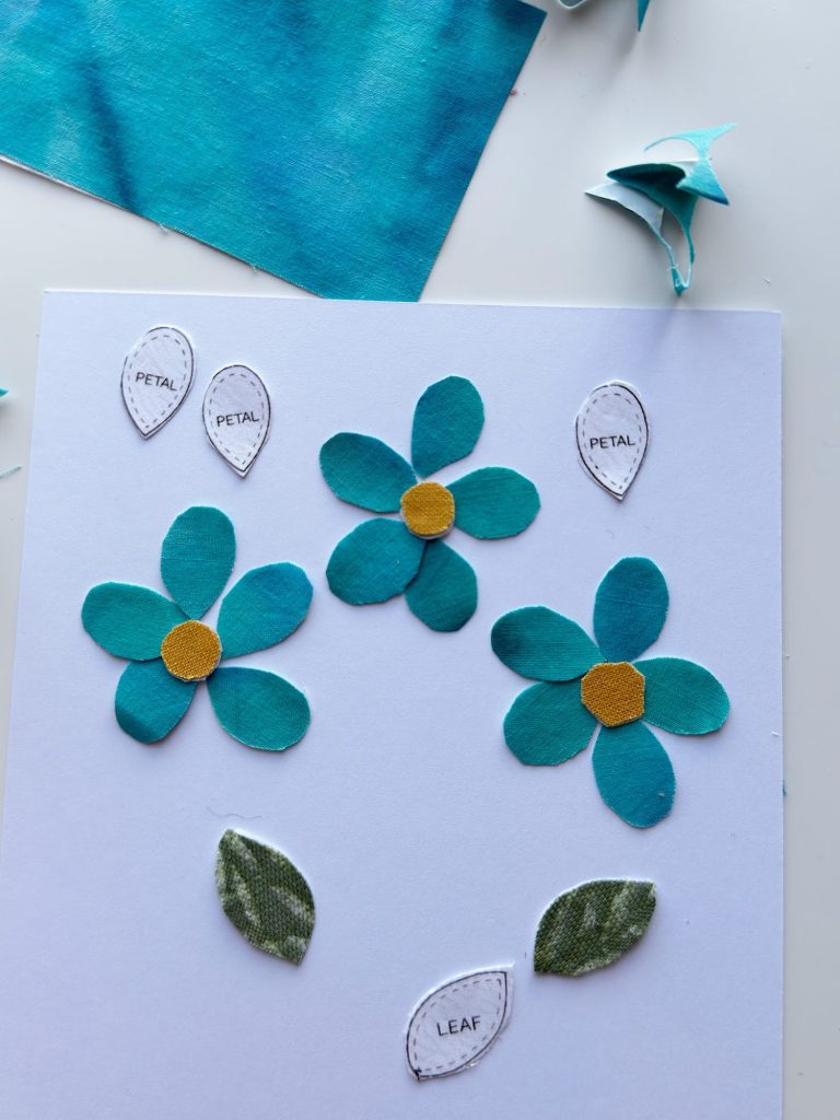 A white paper with three blue fabric flowers, two fabric leaves, and three labeled paper petal and leaf templates arranged on top.